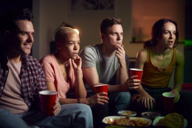 group of friends with drinks sitting on sofa at home watching horror movie together